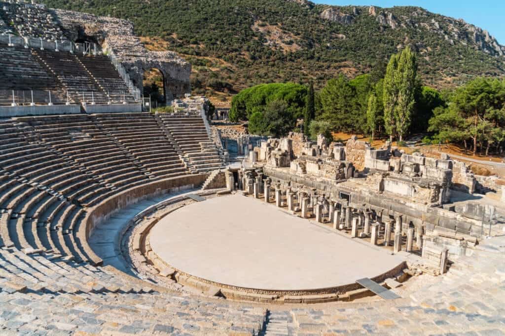 Wide-angle view of the Grand Theatre of Ephesus with stone seating, stage ruins, and forested hills in the background.