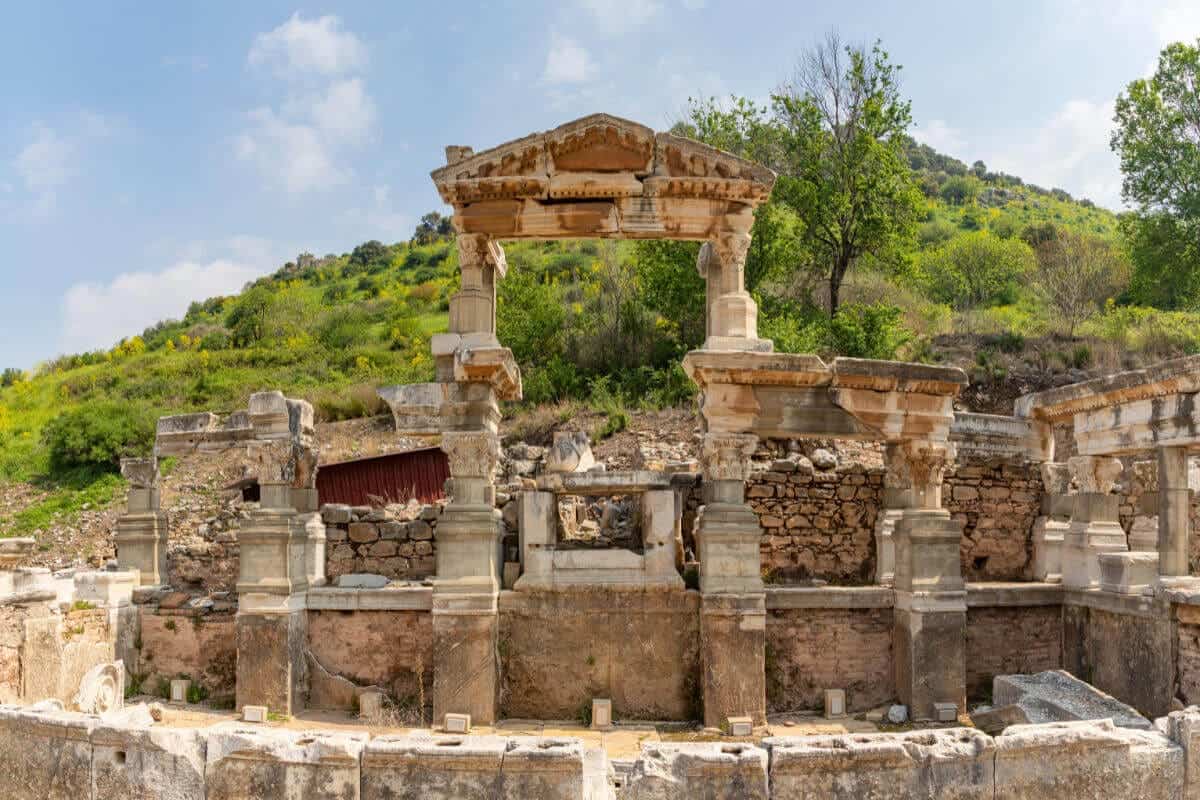 Ruins of the Fountain of Trajan in Ephesus with preserved Roman columns and stone reliefs amid lush hillside.