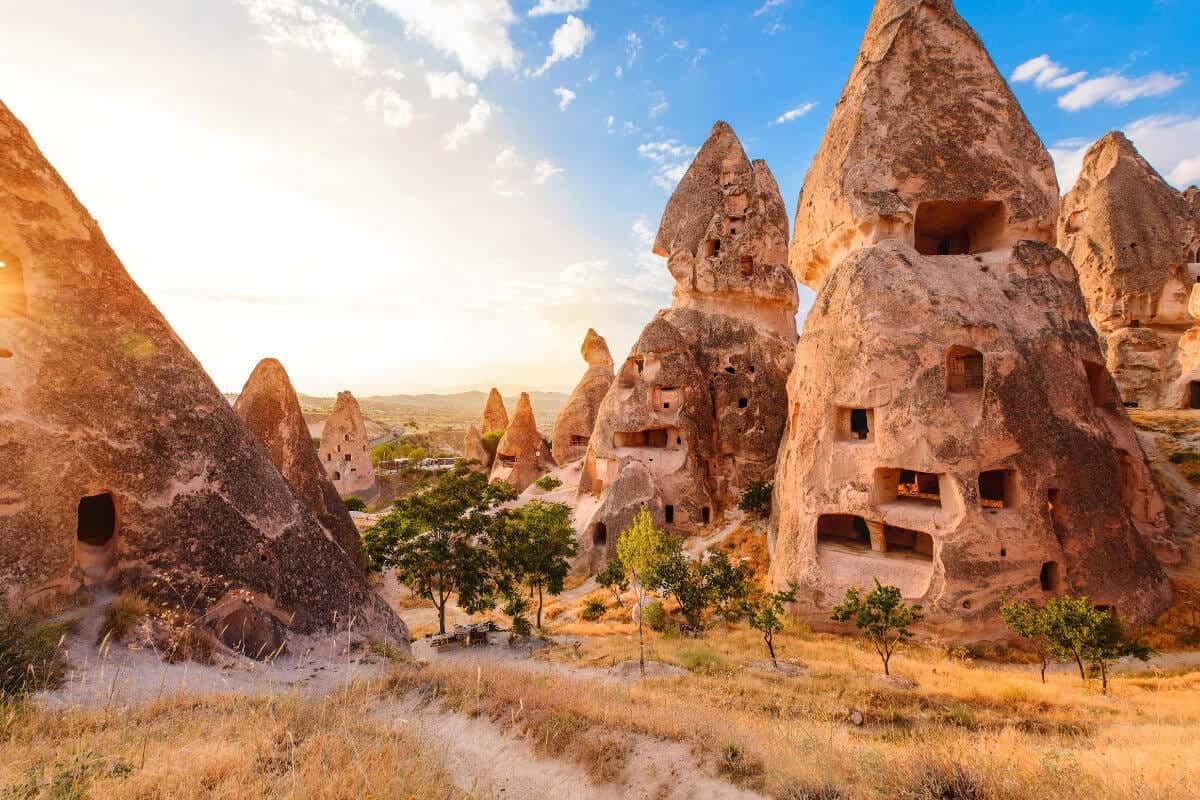 Sunlit fairy chimneys with carved cave dwellings surrounded by dry grass and trees in Cappadocia, Turkey.