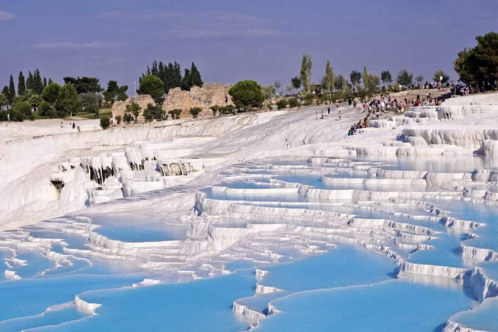 Blue thermal pools cascading down white travertine terraces in Pamukkale, Turkey, with tourists exploring the site.