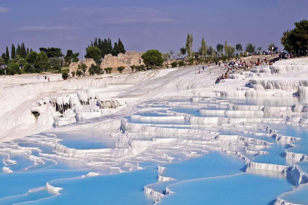 Blue thermal pools cascading down white travertine terraces in Pamukkale, Turkey, with tourists exploring the site.