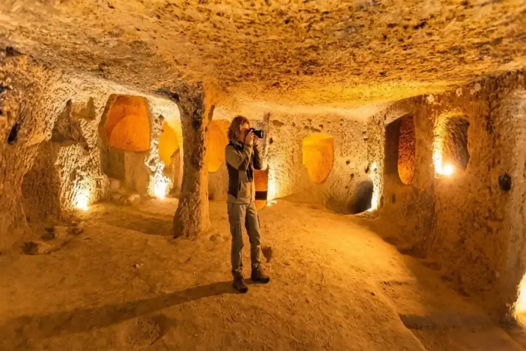 A female tourist with a camera exploring the intricately carved, warmly lit chambers of an ancient underground city, such as Derinkuyu or Kaymakli, in Cappadocia, Turkey.