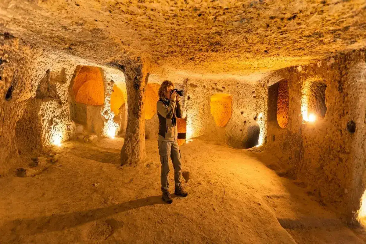A female tourist with a camera exploring the intricately carved, warmly lit chambers of an ancient underground city, such as Derinkuyu or Kaymakli, in Cappadocia, Turkey.