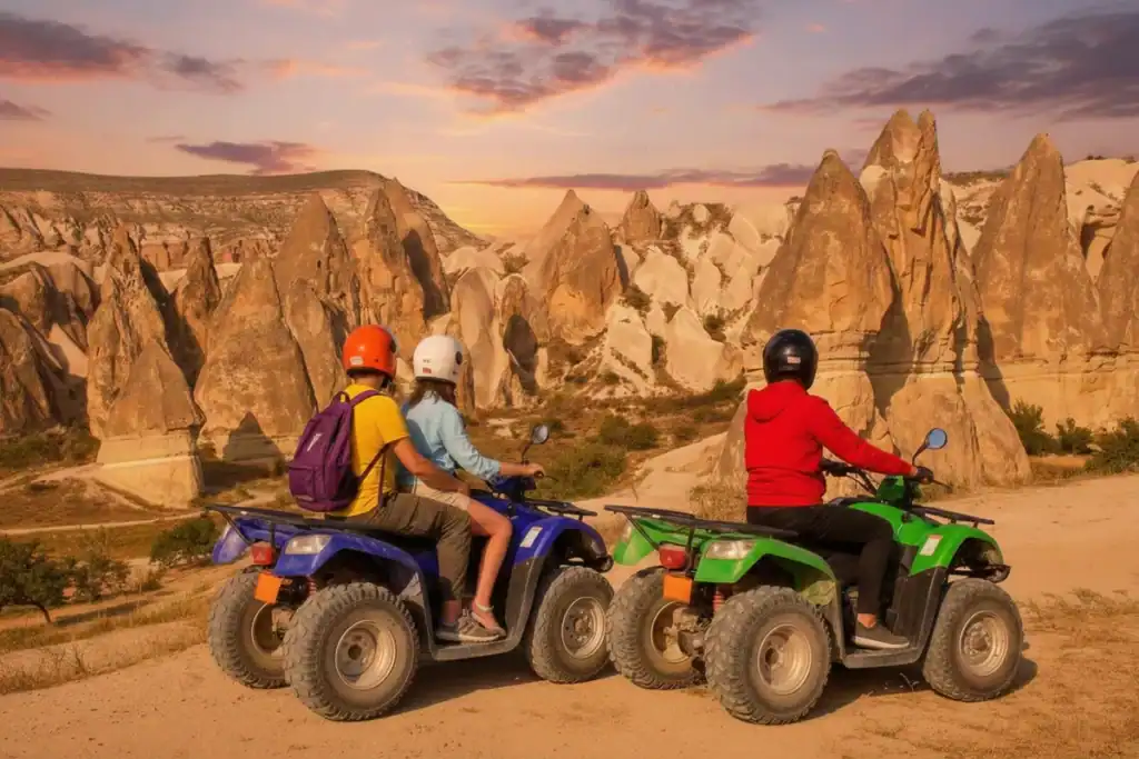 Two riders on a blue ATV and one rider on a green ATV pause on a dirt path to view the unique rock formations of Cappadocia, Turkey, under a dramatic orange and purple sunset sky.