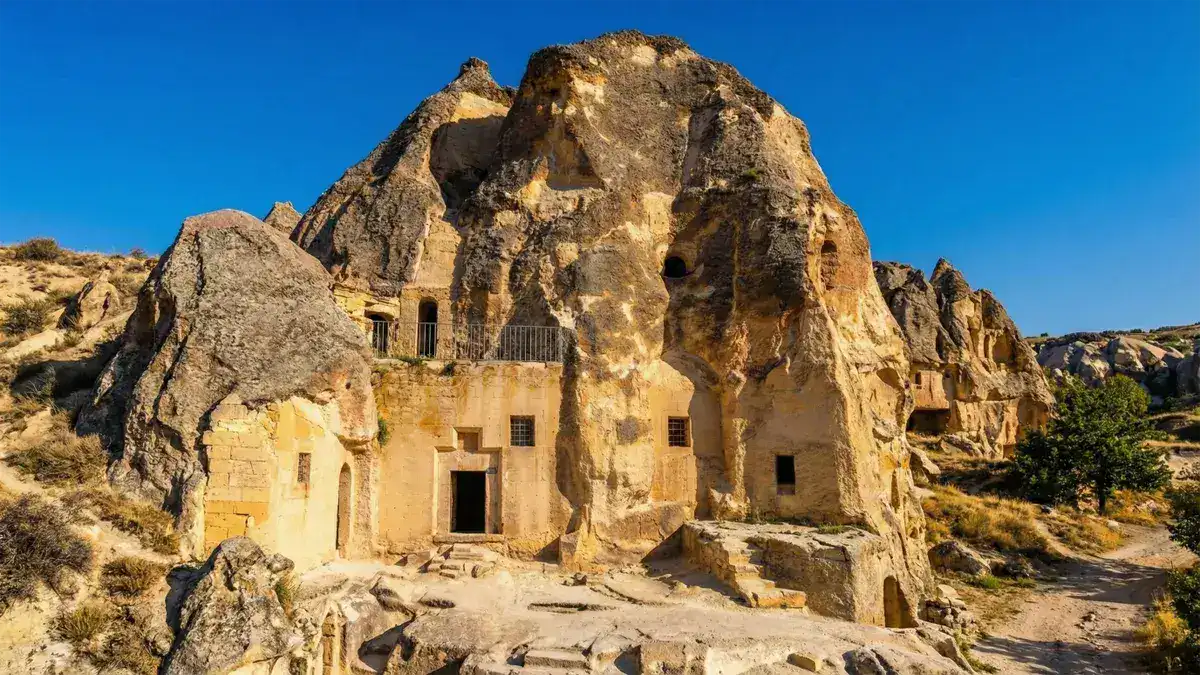 A wide-angle landscape photograph of the ancient rock-cut Keslik Monastery complex in Cappadocia, Turkey, showing carved entrances, windows, stone steps, and a metal railing under a clear deep blue sky.