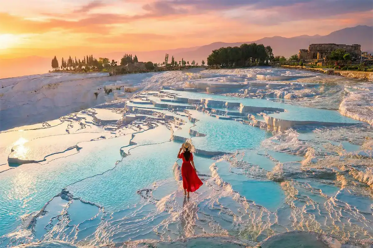 A traveler in a red dress admiring the sunset view over the turquoise thermal pools and white travertine terraces of Pamukkale, a highlight included in the 4 Days Cappadocia and Pamukkale Tour from Istanbul.