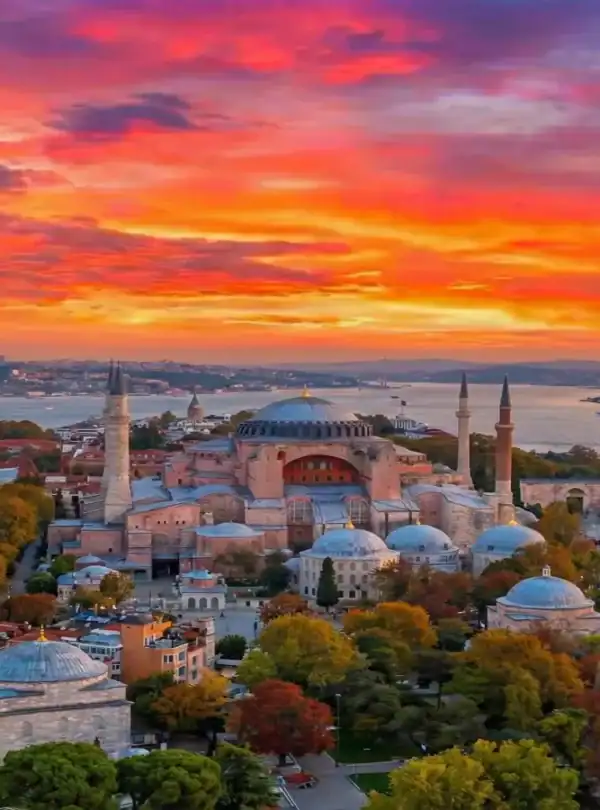 An aerial photograph showing the Hagia Sophia mosque and surrounding buildings in Istanbul, Turkey, at sunset. The sky is filled with dramatic orange, pink, and purple clouds, and the city and Bosphorus Strait are illuminated by the warm light.