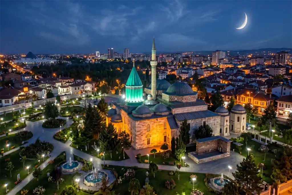 Aerial night view of the illuminated Mevlana Museum in Konya, Turkey, featuring its iconic green dome under a crescent moon and light clouds.