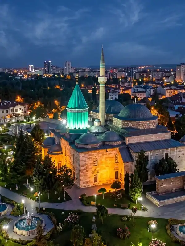 Aerial night view of the illuminated Mevlana Museum in Konya, Turkey, featuring its iconic green dome under a crescent moon and light clouds.
