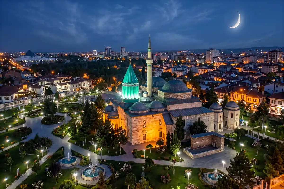 Aerial night view of the illuminated Mevlana Museum in Konya, Turkey, featuring its iconic green dome under a crescent moon and light clouds.