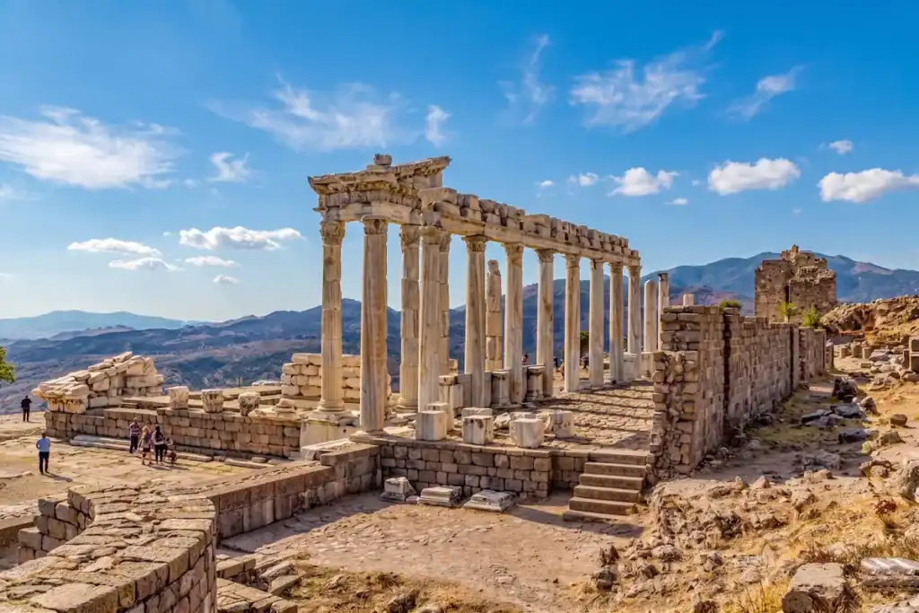 A panoramic view of the ancient Temple of Trajan ruins at Pergamon, Turkey, featuring standing Corinthian columns on a hilltop overlooking a vast scenic valley and other structures.