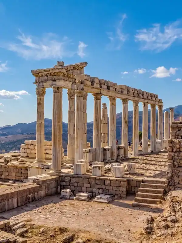 A panoramic view of the ancient Temple of Trajan ruins at Pergamon, Turkey, featuring standing Corinthian columns on a hilltop overlooking a vast scenic valley and other structures.