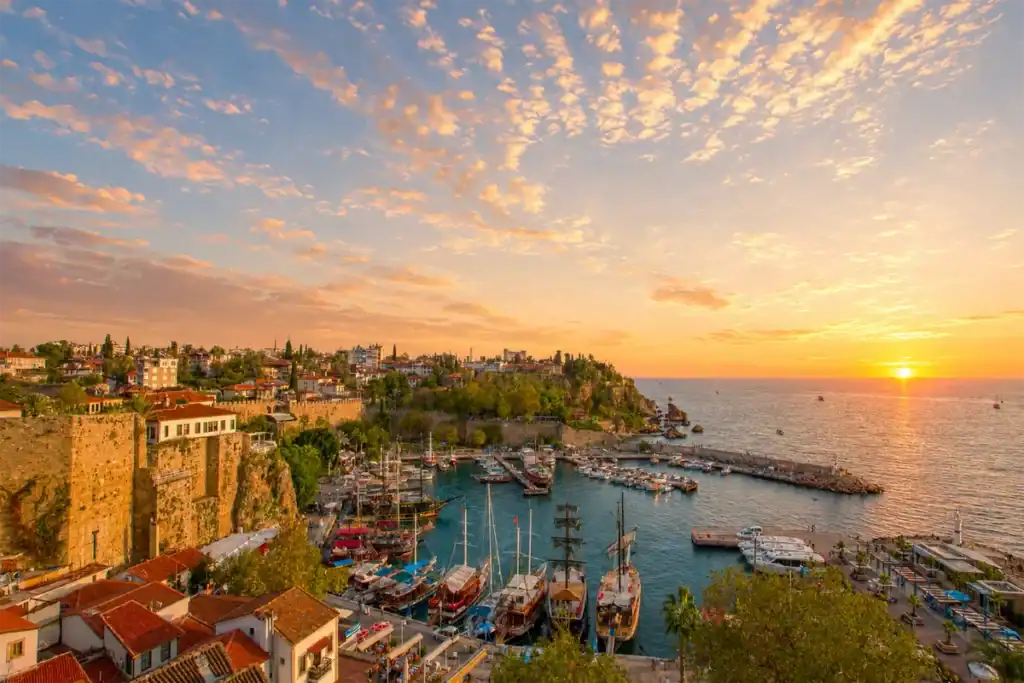 Sunrise over Antalya Kaleici Old Harbor with boats, historic cliffs, red-roofed houses, and light clouds.