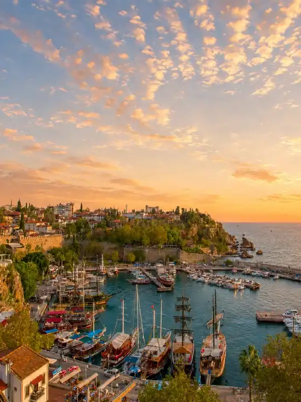 Sunrise over Antalya Kaleici Old Harbor with boats, historic cliffs, red-roofed houses, and light clouds.